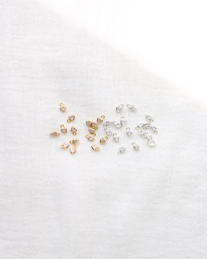Puede incluir: Una colecci&oacute;n de peque&ntilde;os dijes en forma de coraz&oacute;n. Algunos son dorados con piedras transparentes, mientras que otros son plateados con piedras transparentes. Los dijes est&aacute;n dispersos sobre una superficie blanca y texturizada.