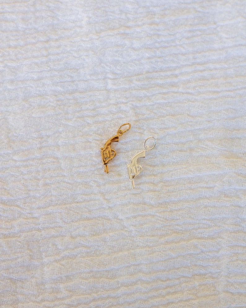 Pode incluir: Dois pingentes em forma de pistola, um dourado e outro prateado, sobre um fundo branco texturizado.
