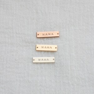 K&ouml;nnte beinhalten: Drei rechteckige Metallanh&auml;nger mit dem Wort "MAMA" eingepr&auml;gt. Die Anh&auml;nger sind in Ros&eacute;gold, Gold und Silber gehalten. Jeder Anh&auml;nger hat an jedem Ende ein kleines Loch.