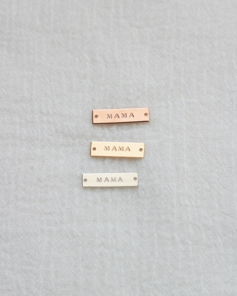 K&ouml;nnte beinhalten: Drei rechteckige Metallanh&auml;nger mit dem Wort "MAMA" eingepr&auml;gt. Die Anh&auml;nger sind in Ros&eacute;gold, Gold und Silber gehalten. Jeder Anh&auml;nger hat an jedem Ende ein kleines Loch.