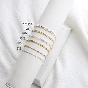 May include: Several fine chain bracelets in gold and silver are displayed on a white cylindrical stand. The bracelets are set against a white fabric background. The image includes text labels: MM183, G46, S46, G19, G13, and S13.