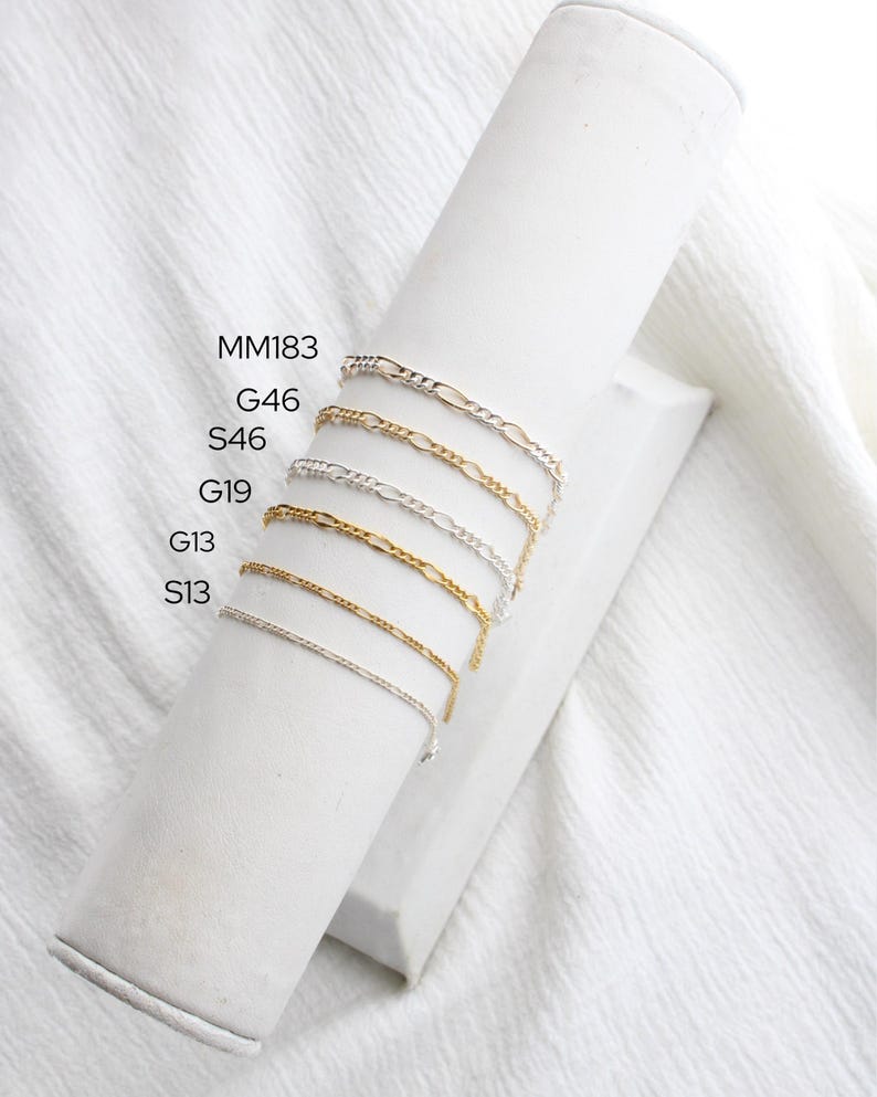 May include: Several fine chain bracelets in gold and silver are displayed on a white cylindrical stand. The bracelets are set against a white fabric background. The image includes text labels: MM183, G46, S46, G19, G13, and S13.