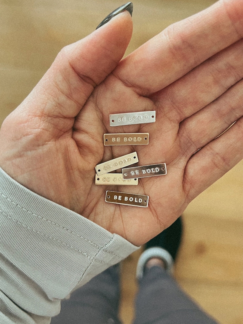K&ouml;nnte beinhalten: Eine Sammlung rechteckiger Metallanh&auml;nger in verschiedenen Metallic-T&ouml;nen, darunter Silber und Gold, die jeweils mit den Worten "BE BOLD" graviert sind. Die Anh&auml;nger werden in einer offenen Hand gehalten, um ihre Gr&ouml;&szlig;e und ihr Design zu zeigen.