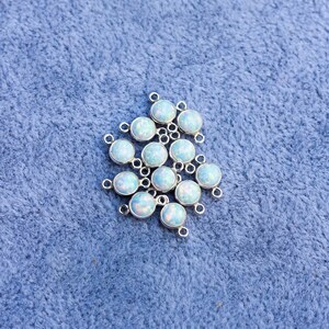 Puede incluir: Un conjunto de conectores plateados, cada uno con una piedra redonda opalescente iridiscente. Las piedras tienen una base blanca lechosa con destellos azules y verdes. Los conectores est&aacute;n sobre una superficie azul texturizada.