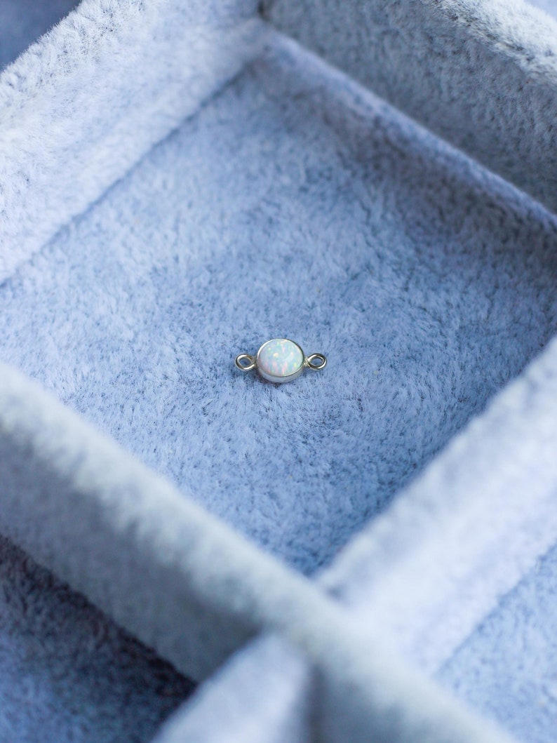 Puede incluir: Un peque&ntilde;o componente de joyer&iacute;a dorado con una piedra de &oacute;palo blanca y redonda en el centro. El &oacute;palo tiene una calidad sutil e iridiscente. El componente tiene peque&ntilde;os bucles a cada lado. El art&iacute;culo est&aacute; sobre un fondo azul claro y suave.