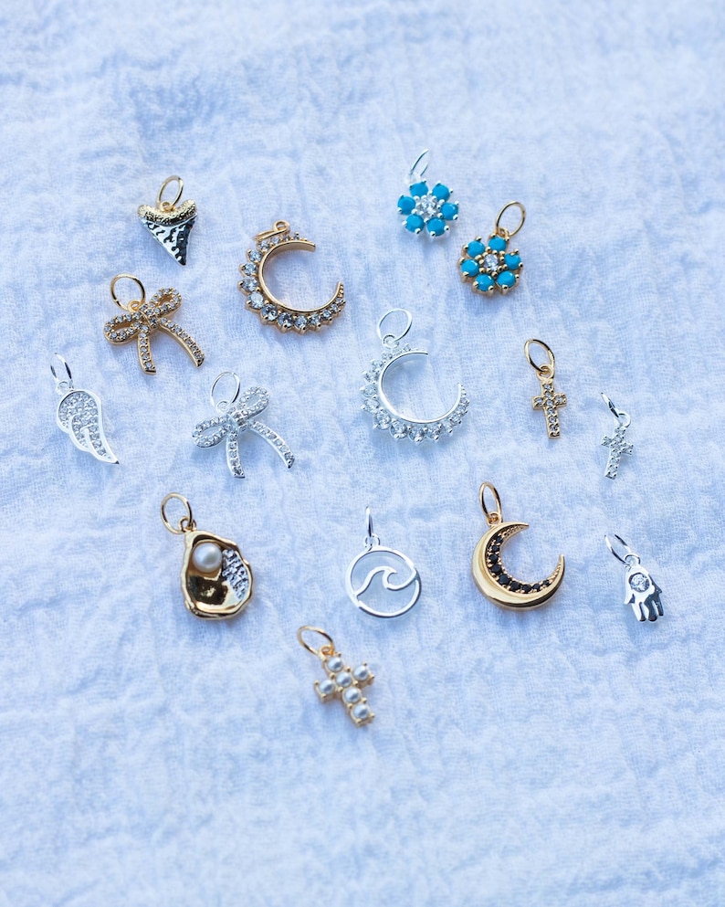 K&ouml;nnte beinhalten: Eine Sammlung von 14 silbernen und goldenen Anh&auml;ngern mit verschiedenen Designs, darunter ein Halbmond, ein Kreuz, eine Welle, eine Hand, eine Schleife, eine Muschel, eine Haifischflosse und eine Blume. Die Anh&auml;nger sind auf einem wei&szlig;en Stoffhintergrund angeordnet.