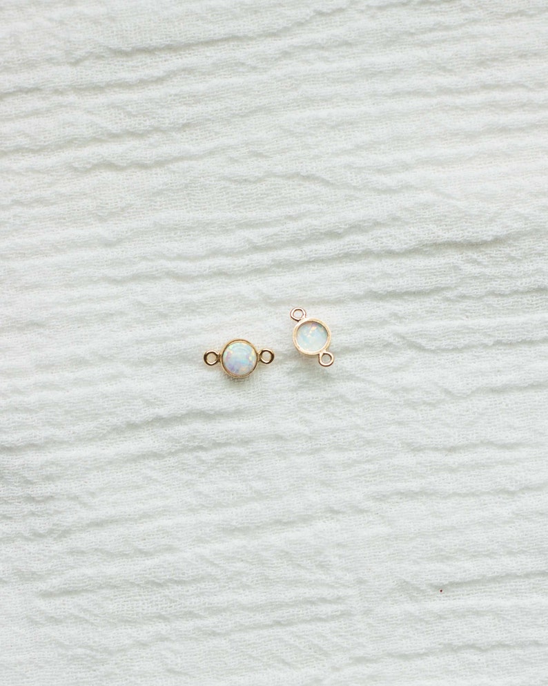 Puede incluir: Dos peque&ntilde;os conectores de joyer&iacute;a dorados, cada uno con una piedra redonda iridiscente similar al &oacute;palo. Los conectores tienen peque&ntilde;os bucles a cada lado para sujetarlos a otros componentes de joyer&iacute;a. El fondo es una tela blanca texturizada.