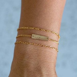 K&ouml;nnte beinhalten: Drei goldene Armb&auml;nder sind an einem Handgelenk gestapelt. Die oberen und unteren Armb&auml;nder haben zarte Kettenglieder. Das mittlere Armband hat einen rechteckigen Balken mit dem eingravierten Wort "MAMA". Die Armb&auml;nder sind warmgoldfarben.