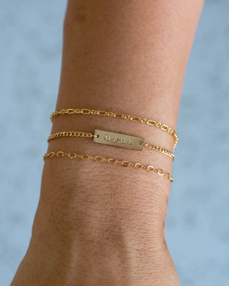 K&ouml;nnte beinhalten: Drei goldene Armb&auml;nder sind an einem Handgelenk gestapelt. Die oberen und unteren Armb&auml;nder haben zarte Kettenglieder. Das mittlere Armband hat einen rechteckigen Balken mit dem eingravierten Wort "MAMA". Die Armb&auml;nder sind warmgoldfarben.