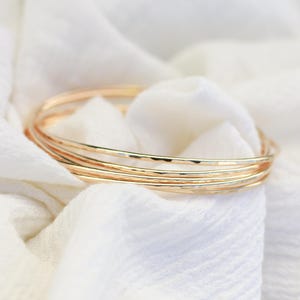 May include: A stack of gold-colored bangles rests on a white, textured fabric. The bangles have a hammered texture, reflecting light. The jewelry is simple and elegant, suitable for everyday wear.