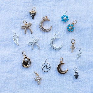 K&ouml;nnte beinhalten: Eine Sammlung von 14 silbernen und goldenen Charms mit verschiedenen Designs, darunter eine Mondsichel, ein Kreuz, eine Welle, ein Herz, eine Schleife, eine Blume, eine Hand und eine Austernschalen. Die Charms sind auf einem wei&szlig;en Stoffhintergrund angeordnet.