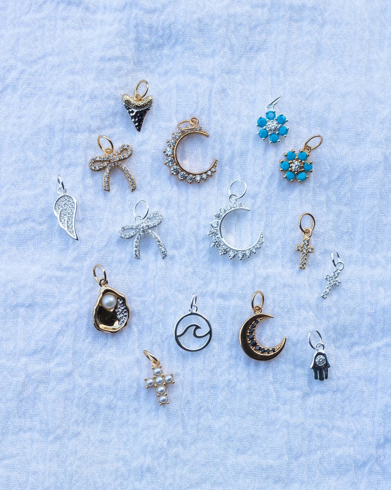 K&ouml;nnte beinhalten: Eine Sammlung von 14 silbernen und goldenen Charms mit verschiedenen Designs, darunter eine Mondsichel, ein Kreuz, eine Welle, ein Herz, eine Schleife, eine Blume, eine Hand und eine Austernschalen. Die Charms sind auf einem wei&szlig;en Stoffhintergrund angeordnet.