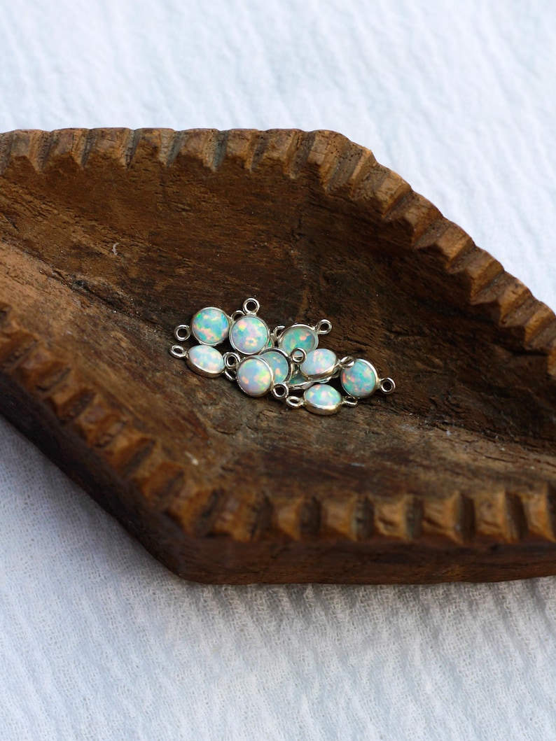 Puede incluir: Un conjunto de peque&ntilde;os colgantes de &oacute;palo ovalados con engastes plateados. Los &oacute;palos tienen una base blanca lechosa con destellos iridiscentes azules y verdes. Los colgantes est&aacute;n dispersos sobre una superficie de madera oscura.