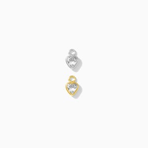 Puede incluir: Dos colgantes en forma de coraz&oacute;n, uno plateado y otro dorado, cada uno con un centro de piedra transparente y brillante. Los colgantes tienen un peque&ntilde;o lazo en la parte superior para sujetarlos a un collar o pulsera. El fondo es blanco.