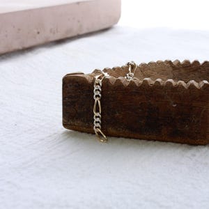 May include: A gold and silver chain bracelet with a Figaro link design. The bracelet is displayed on a wooden object with a textured surface, set against a white background. The bracelet's links alternate between gold and silver.