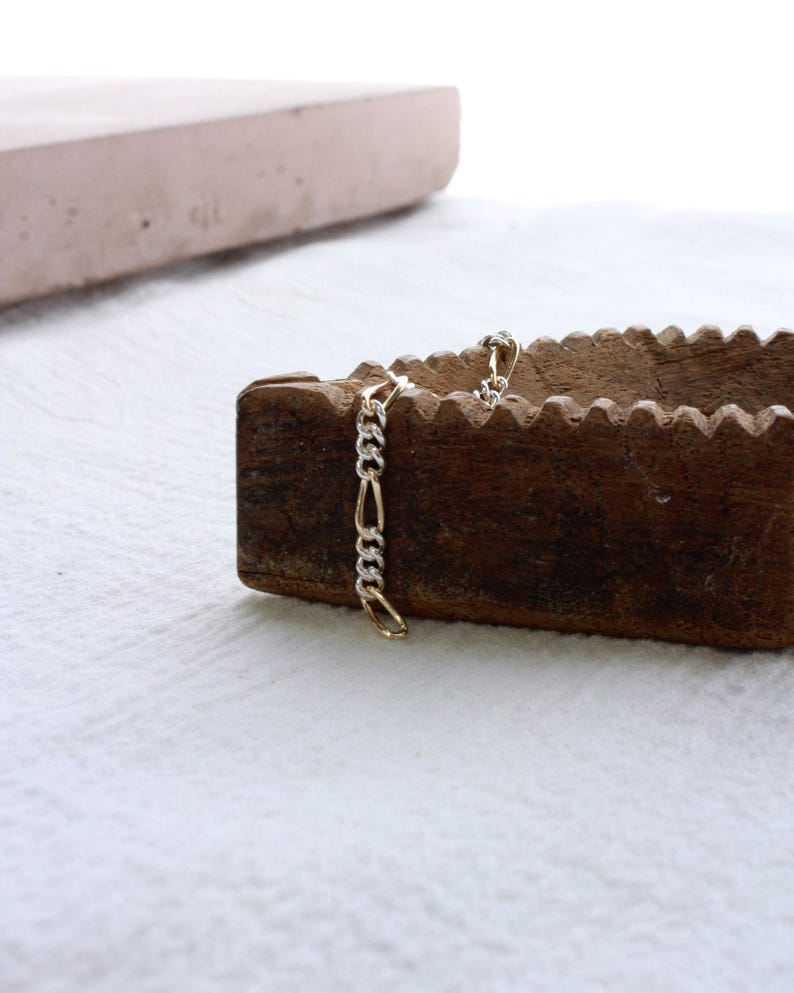 May include: A gold and silver chain bracelet with a Figaro link design. The bracelet is displayed on a wooden object with a textured surface, set against a white background. The bracelet's links alternate between gold and silver.