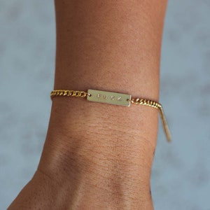 K&ouml;nnte beinhalten: Goldfarbenes Kettenarmband mit einem rechteckigen Steg mit der Aufschrift "FUCK". Das Armband wird am Handgelenk getragen. Die Kette hat ein Panzerketten-Design.