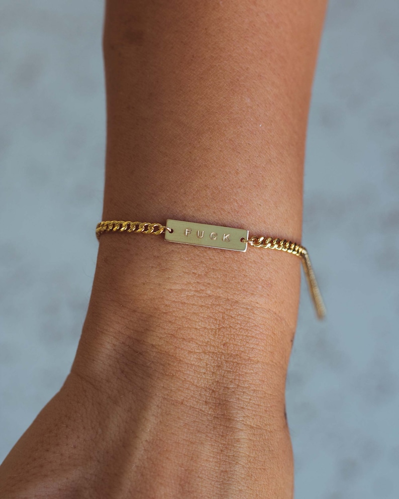 K&ouml;nnte beinhalten: Goldfarbenes Kettenarmband mit einem rechteckigen Steg mit der Aufschrift "FUCK". Das Armband wird am Handgelenk getragen. Die Kette hat ein Panzerketten-Design.