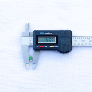 May include: A digital caliper measuring a small green gemstone pendant. The caliper reads 4.55 mm.