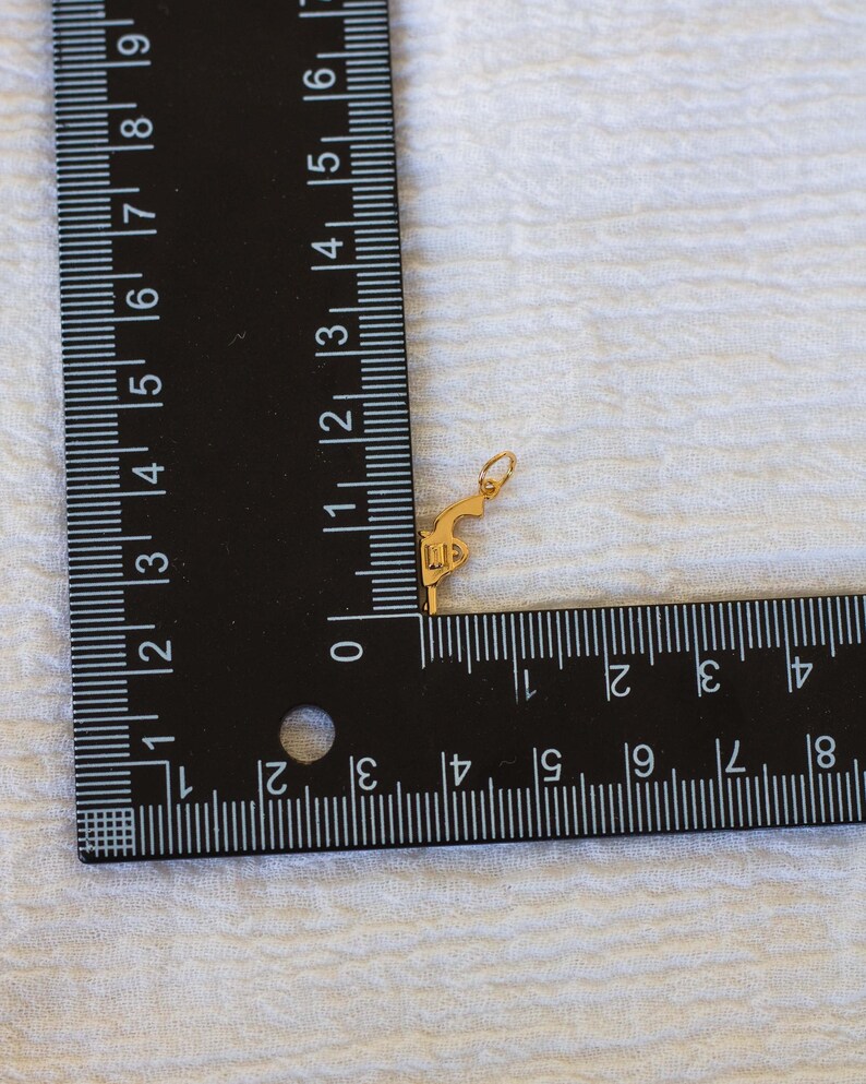 Pode incluir: Um amuleto dourado em forma de pistola com as letras "11" gravadas no lado. O amuleto &eacute; medido com uma r&eacute;gua.