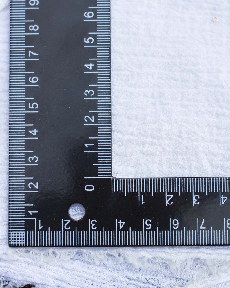 May include: A black ruler with white markings is placed on a white fabric surface. The ruler is marked in inches and centimeters. The ruler is positioned at a 90-degree angle.