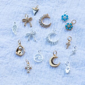 K&ouml;nnte beinhalten: Eine Sammlung von 15 silbernen und goldenen Anh&auml;ngern mit verschiedenen Designs, darunter ein Halbmond, ein Kreuz, eine Welle, eine Muschel, eine Schleife, ein Stern, eine Hand und eine Haifischflosse. Die Anh&auml;nger sind alle mit funkelnden Kristallen verziert.
