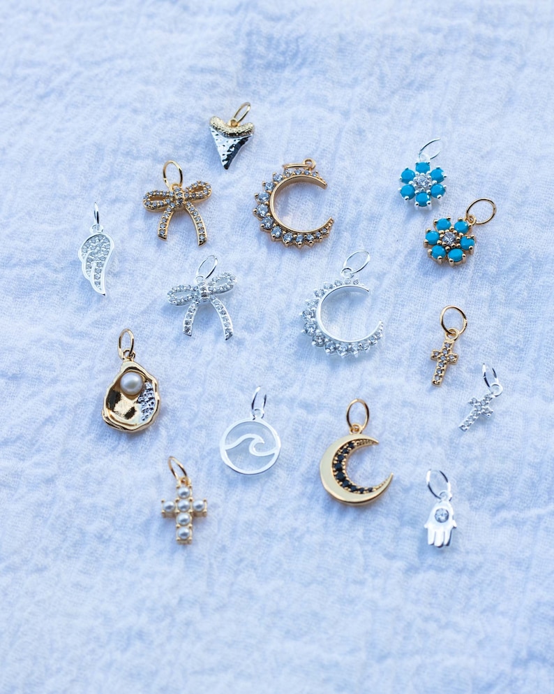 K&ouml;nnte beinhalten: Eine Sammlung von 15 silbernen und goldenen Anh&auml;ngern mit verschiedenen Designs, darunter ein Halbmond, ein Kreuz, eine Welle, eine Muschel, eine Schleife, ein Stern, eine Hand und eine Haifischflosse. Die Anh&auml;nger sind alle mit funkelnden Kristallen verziert.