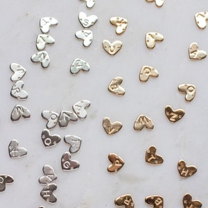 May include: A collection of gold and silver heart-shaped charms with different letters engraved on them. The charms are arranged on a white surface.