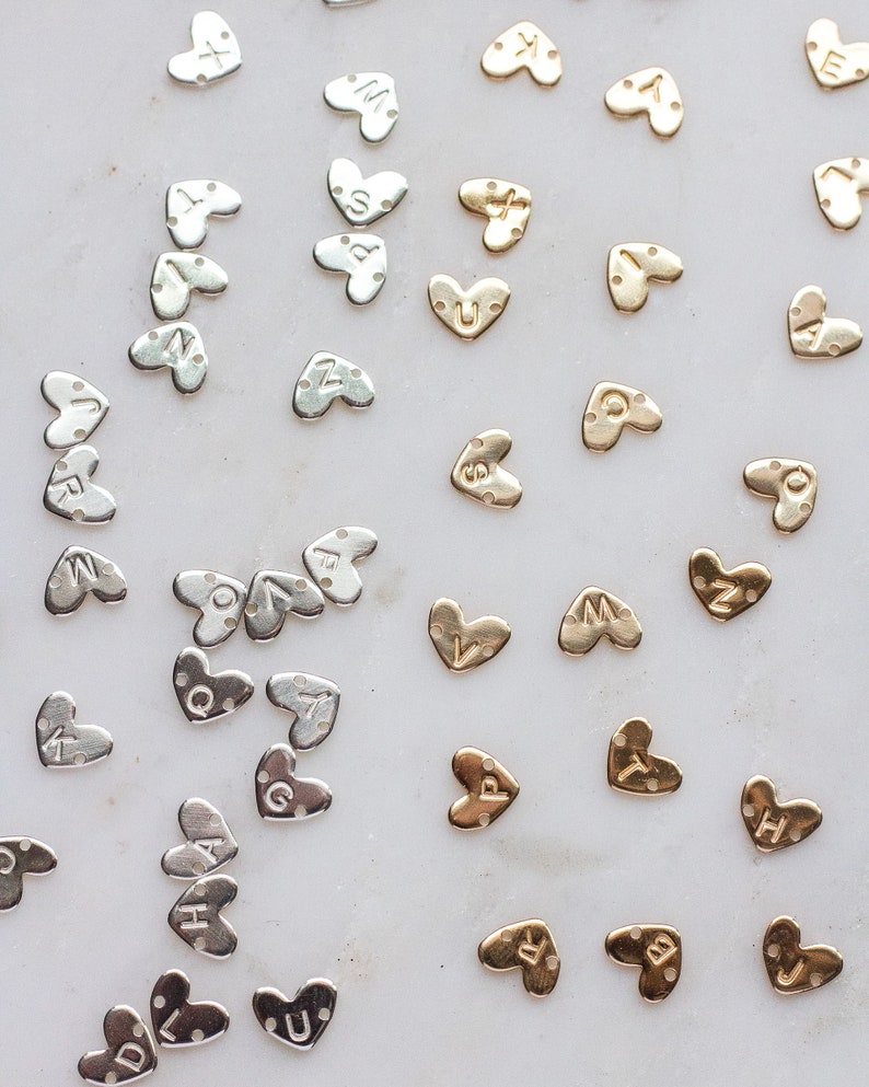 May include: A collection of gold and silver heart-shaped charms with different letters engraved on them. The charms are arranged on a white surface.