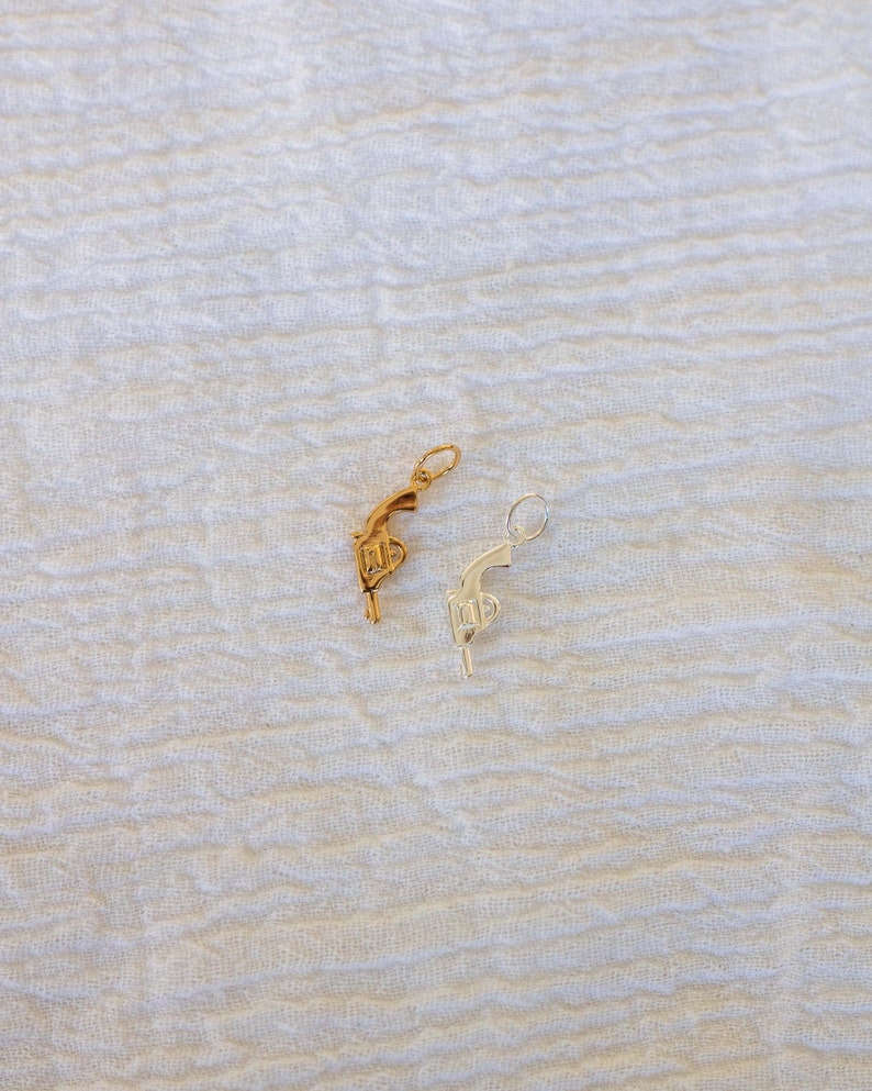 Pode incluir: Dois pingentes em forma de pistola, um dourado e outro prateado, sobre um fundo branco texturizado.