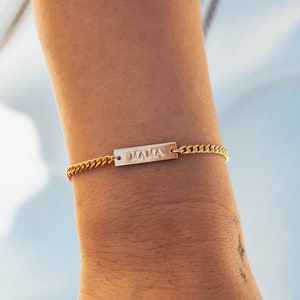 K&ouml;nnte beinhalten: Goldfarbenes Armband mit einem rechteckigen Anh&auml;nger mit der Aufschrift "MAMA". Das Armband hat ein Panzerketten-Design. Das Bild zeigt das Armband an einem Handgelenk.