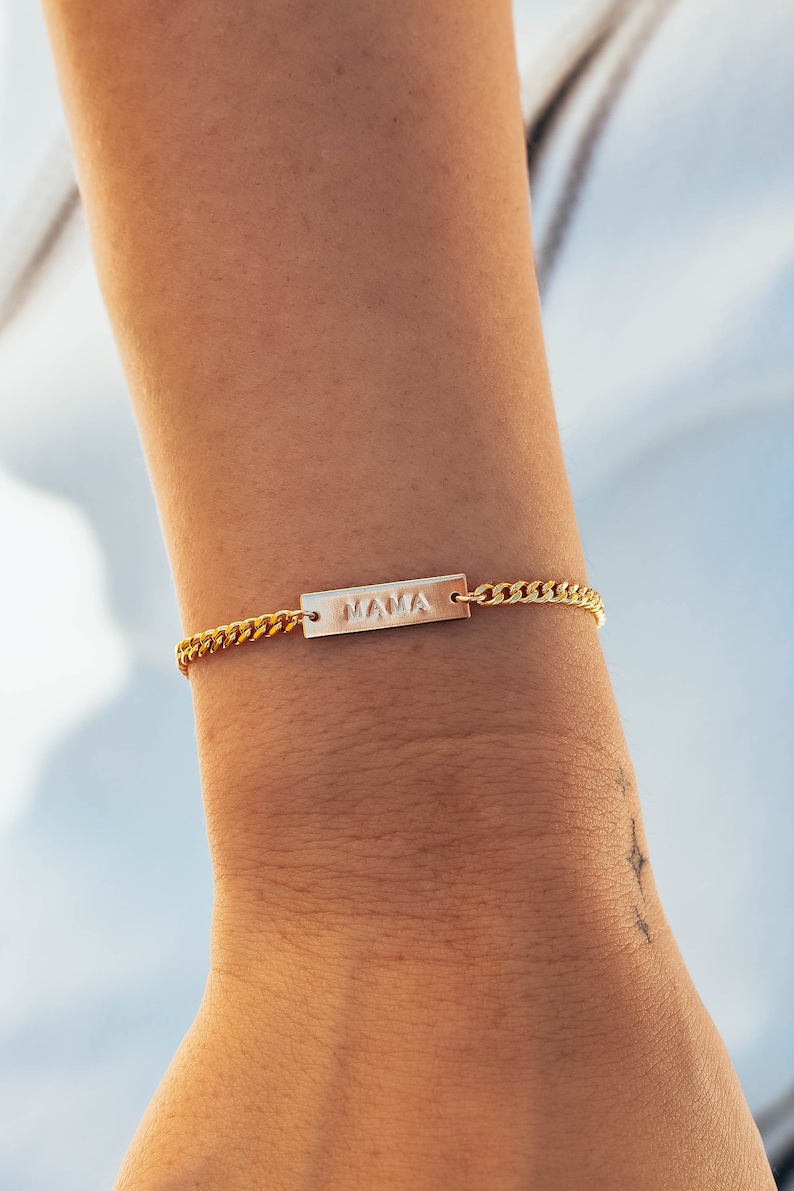 K&ouml;nnte beinhalten: Goldfarbenes Armband mit einem rechteckigen Anh&auml;nger mit der Aufschrift "MAMA". Das Armband hat ein Panzerketten-Design. Das Bild zeigt das Armband an einem Handgelenk.