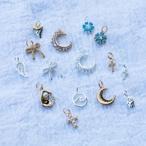 K&ouml;nnte beinhalten: Eine Sammlung von Silber- und Goldanh&auml;ngern mit verschiedenen Designs, darunter eine Mondsichel, ein Kreuz, eine Welle, eine Muschel, eine Hand, eine Schleife, eine Blume und ein Engelsfl&uuml;gel. Die Anh&auml;nger sind auf einem wei&szlig;en Stoffhintergrund angeordnet.