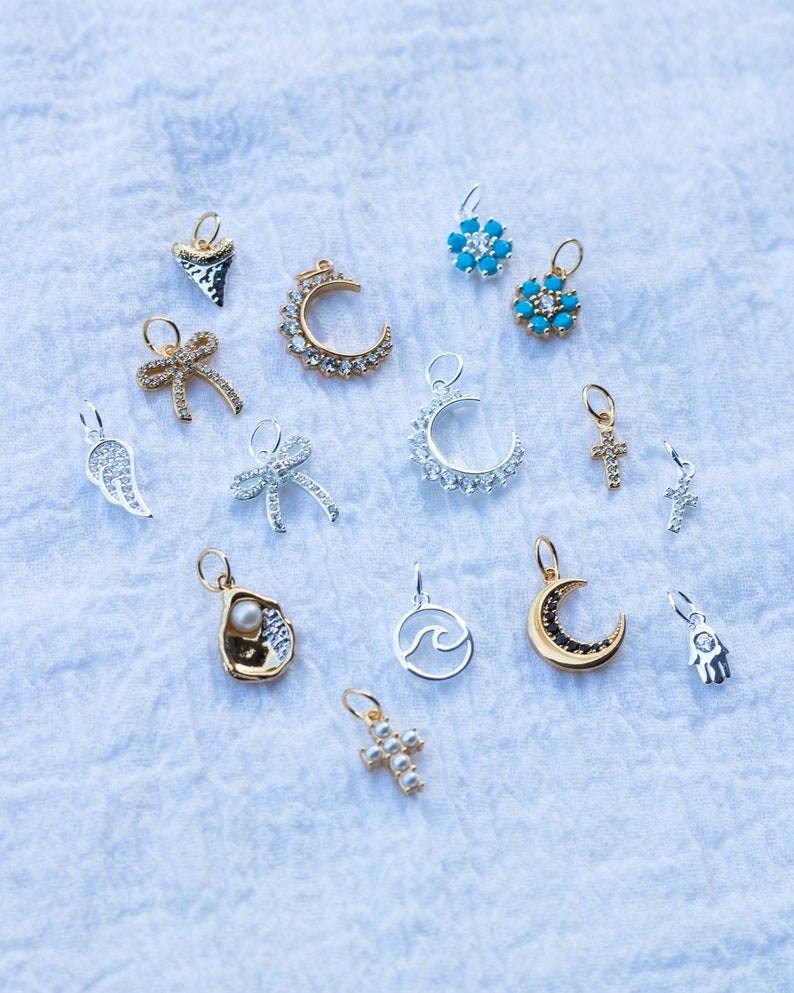 K&ouml;nnte beinhalten: Eine Sammlung von Silber- und Goldanh&auml;ngern mit verschiedenen Designs, darunter eine Mondsichel, ein Kreuz, eine Welle, eine Muschel, eine Hand, eine Schleife, eine Blume und ein Engelsfl&uuml;gel. Die Anh&auml;nger sind auf einem wei&szlig;en Stoffhintergrund angeordnet.