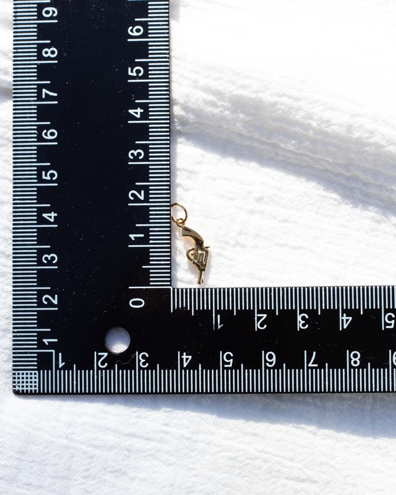 Pode incluir: Um amuleto dourado em forma de pistola, medido com uma r&eacute;gua. O amuleto mede aproximadamente 3,8 cm de comprimento.