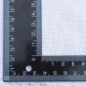 May include: A black ruler with white markings is placed on a white fabric surface. The ruler is marked in inches and centimeters. The ruler is positioned at a 90-degree angle.