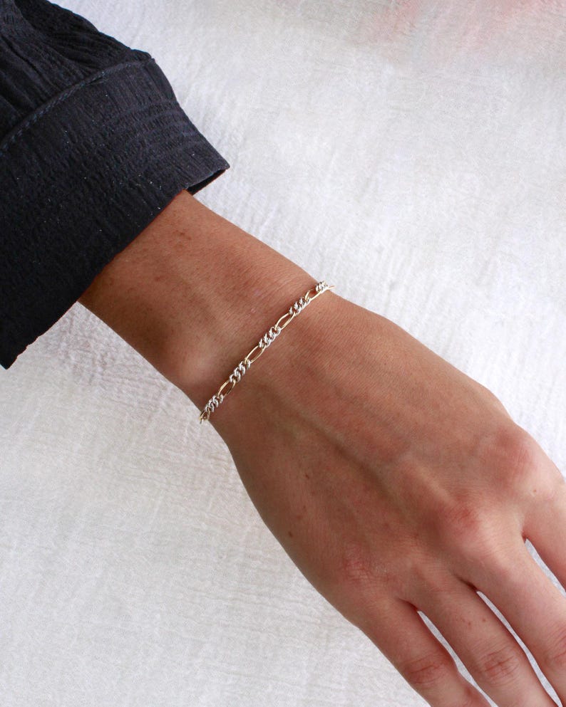 May include: A silver and gold Figaro chain bracelet is shown on a wrist. The bracelet has a pattern of three small links followed by one elongated link. The person is wearing a black long-sleeved shirt.