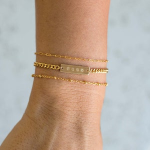 K&ouml;nnte beinhalten: Drei goldene Armb&auml;nder auf einem Handgelenk. Das obere Armband ist eine zarte Kette, das mittlere eine Panzerkette mit einem rechteckigen Balken mit der Aufschrift "FUCK", und das untere Armband ist eine Perlenkette.