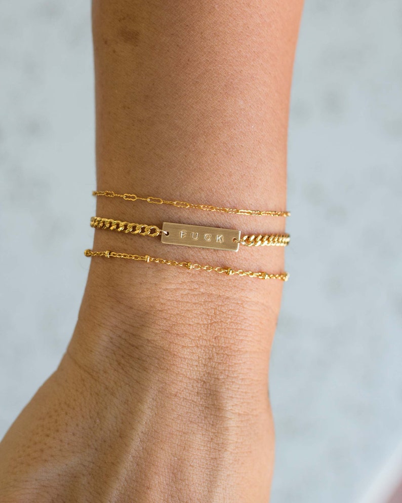 K&ouml;nnte beinhalten: Drei goldene Armb&auml;nder auf einem Handgelenk. Das obere Armband ist eine zarte Kette, das mittlere eine Panzerkette mit einem rechteckigen Balken mit der Aufschrift "FUCK", und das untere Armband ist eine Perlenkette.