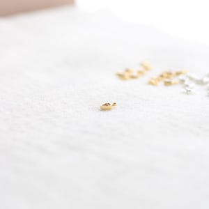Puede incluir: Un peque&ntilde;o dije dorado en forma de coraz&oacute;n, sobre una superficie blanca texturizada. Varios otros dijes similares en oro y plata est&aacute;n ligeramente desenfocados en el fondo. El dije es probablemente para la fabricaci&oacute;n de joyas.