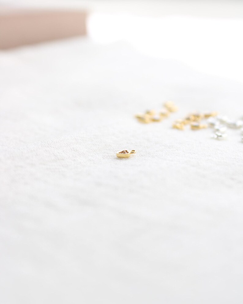Puede incluir: Un peque&ntilde;o dije dorado en forma de coraz&oacute;n, sobre una superficie blanca texturizada. Varios otros dijes similares en oro y plata est&aacute;n ligeramente desenfocados en el fondo. El dije es probablemente para la fabricaci&oacute;n de joyas.