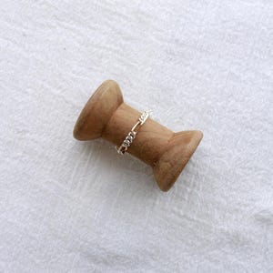 May include: A gold and silver chain ring sits on a wooden spool. The ring has a delicate chain design. The spool is made of light brown wood and is set against a white, textured background.
