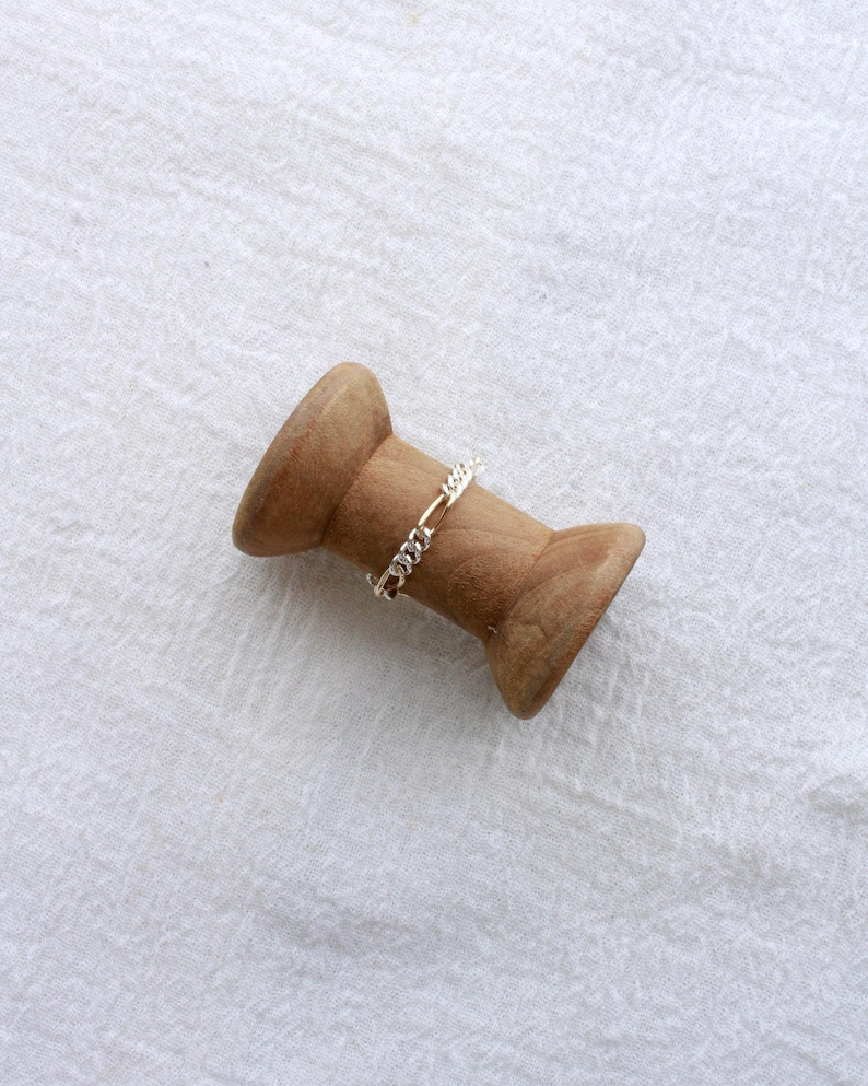 May include: A gold and silver chain ring sits on a wooden spool. The ring has a delicate chain design. The spool is made of light brown wood and is set against a white, textured background.