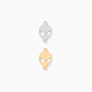 May include: Two alien-shaped charms. One is silver-colored, the other is gold-colored. Each charm has two eye-shaped cutouts and a loop at the top and bottom for attaching to jewelry. The charms are on a white background.