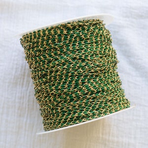 Puede incluir: Un carrete de cadena dorada con un revestimiento de esmalte verde. La cadena está enrollada alrededor de un carrete de plástico blanco.