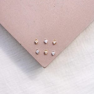 Puede incluir: Seis peque&ntilde;os colgantes en forma de coraz&oacute;n, algunos dorados y otros plateados, cada uno con una piedra transparente y brillante. Los colgantes est&aacute;n dispuestos sobre una superficie marr&oacute;n rosada, creando una exhibici&oacute;n de joyer&iacute;a minimalista. Los colgantes son probablemente para collares o pendientes.
