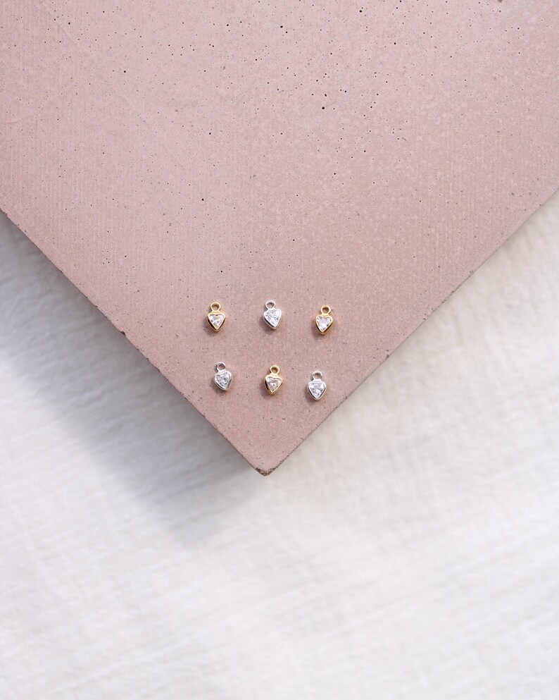Puede incluir: Seis peque&ntilde;os colgantes en forma de coraz&oacute;n, algunos dorados y otros plateados, cada uno con una piedra transparente y brillante. Los colgantes est&aacute;n dispuestos sobre una superficie marr&oacute;n rosada, creando una exhibici&oacute;n de joyer&iacute;a minimalista. Los colgantes son probablemente para collares o pendientes.