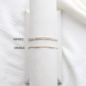 May include: Two delicate bracelets displayed on a white stand. The top bracelet is a gold and silver Figaro chain. The bottom bracelet is gold with small, evenly spaced beads. The text "MM183" and "MM184" are visible.