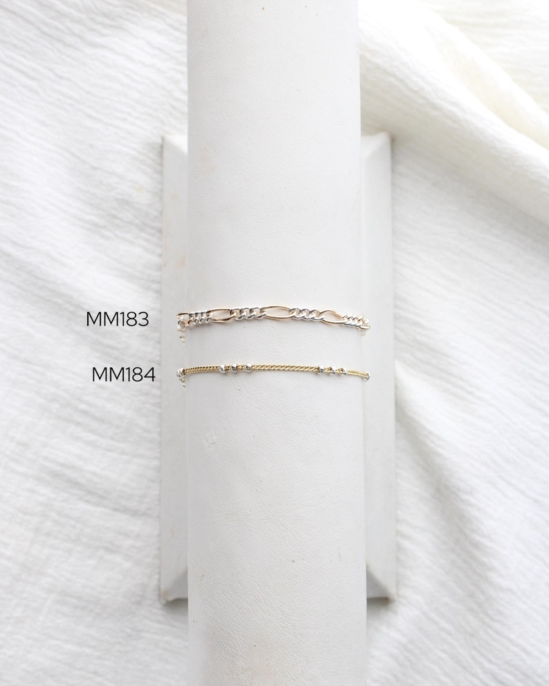 May include: Two delicate bracelets displayed on a white stand. The top bracelet is a gold and silver Figaro chain. The bottom bracelet is gold with small, evenly spaced beads. The text "MM183" and "MM184" are visible.