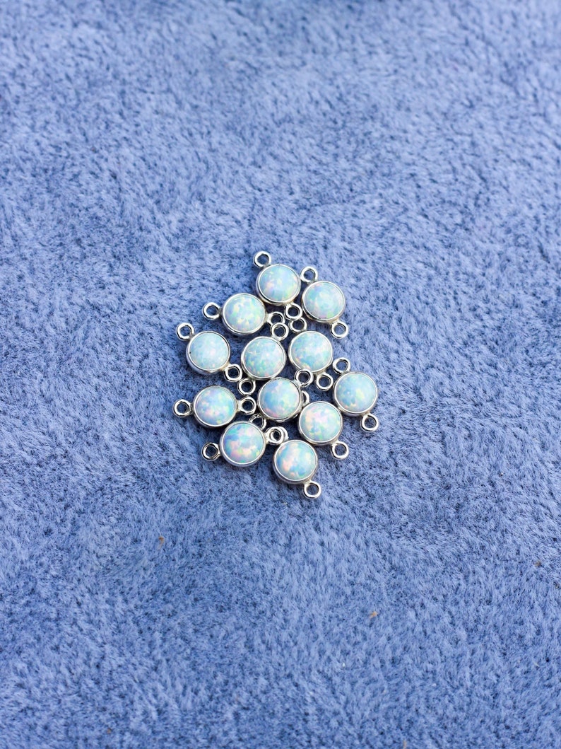 Puede incluir: Un conjunto de componentes de joyer&iacute;a redondos, plateados, con cabujones iridiscentes y opalinos. Cada componente tiene un peque&ntilde;o lazo para la fijaci&oacute;n. Los componentes est&aacute;n dispersos sobre una superficie azul texturizada.